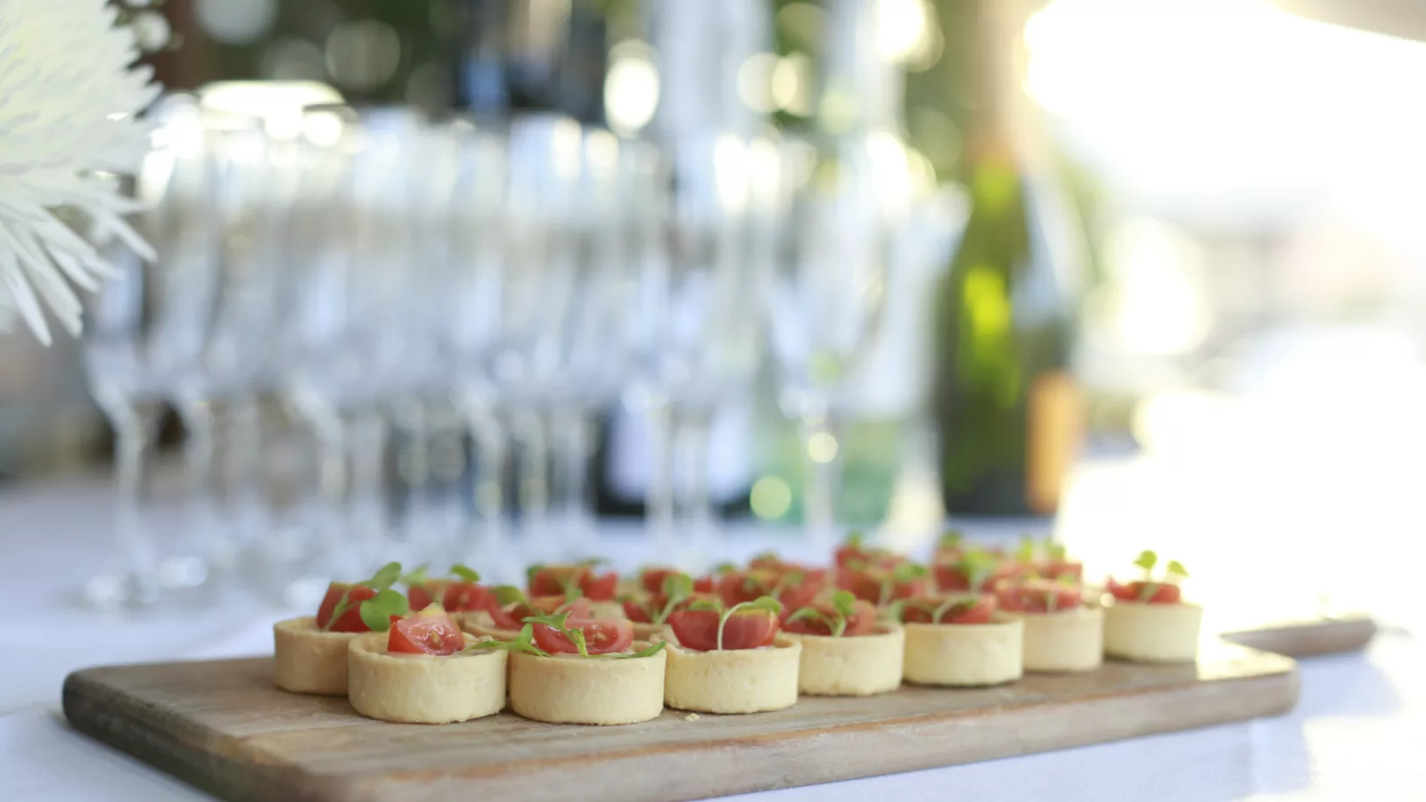 Selection of mini quiches served on a wooden board on a table with a white table cloth and champagne flutes in the background