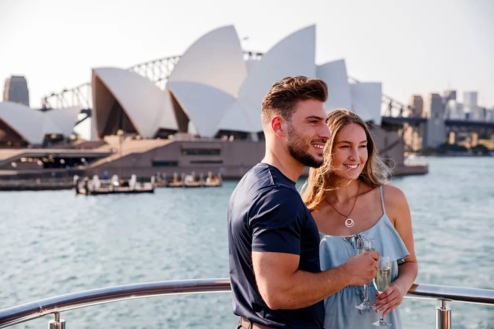 Couple smiling having champagne drinks open deck harbour experience sightseeing with Opera House in the background