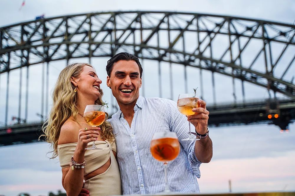 Couple smiling with drinks in front of Harbour Bridge
