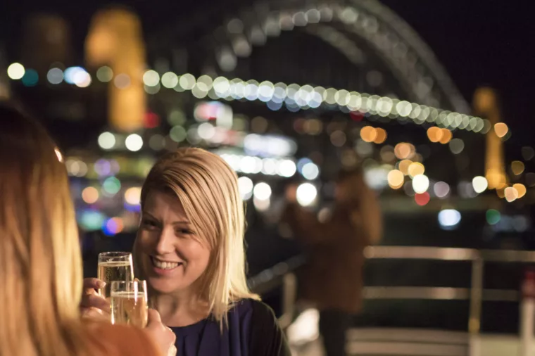 Friends girls having champagne on open deck with Harbour Bridge night lights in the background special events