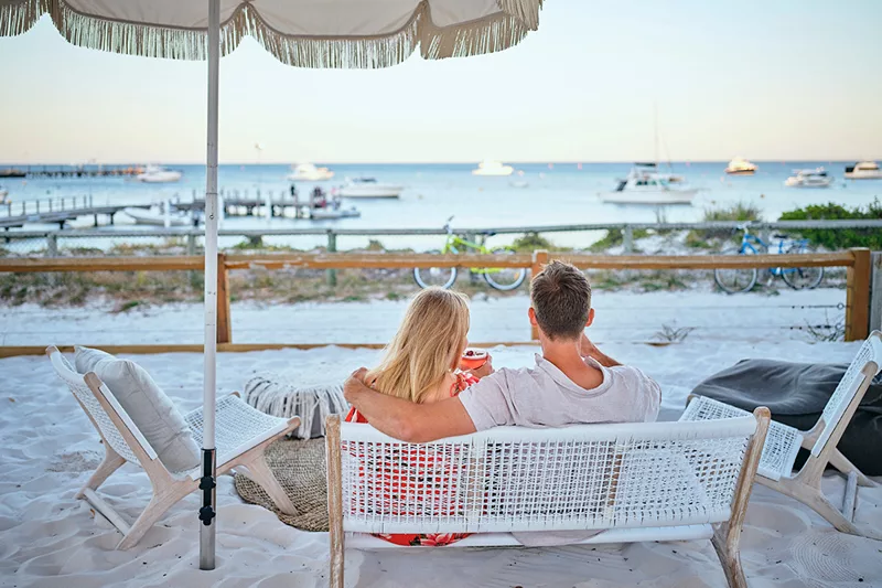Couple sitting on beach lounge chairs enjoying drinks overlooking Thomson Bay at Samphire Rottnest