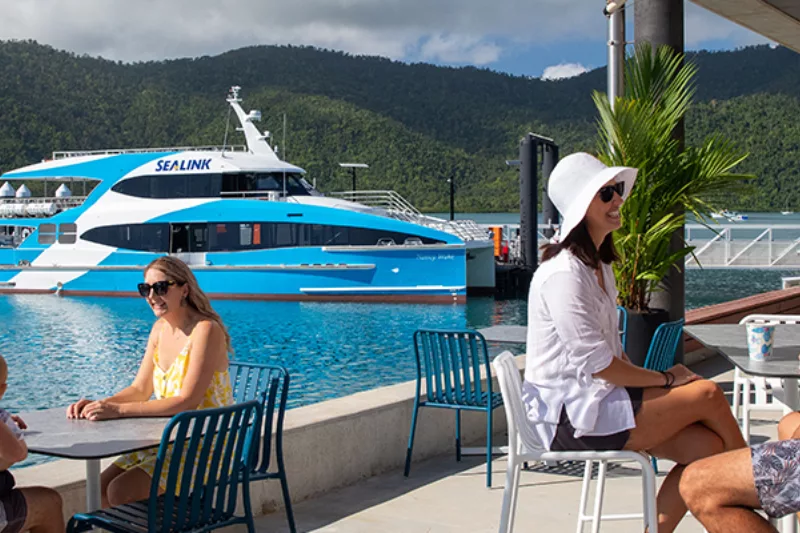 Tourists relax at a cafe next to the harbour, against a blue and white ferry in the water