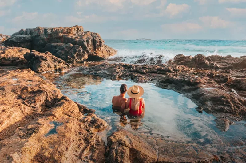A couple swim in a rock pool as they look on at the ocean and the horizon