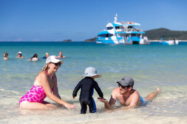 A family of three seen enjoying on the Whitehaven beach with the SeaLink Whitsundays ferry in the background