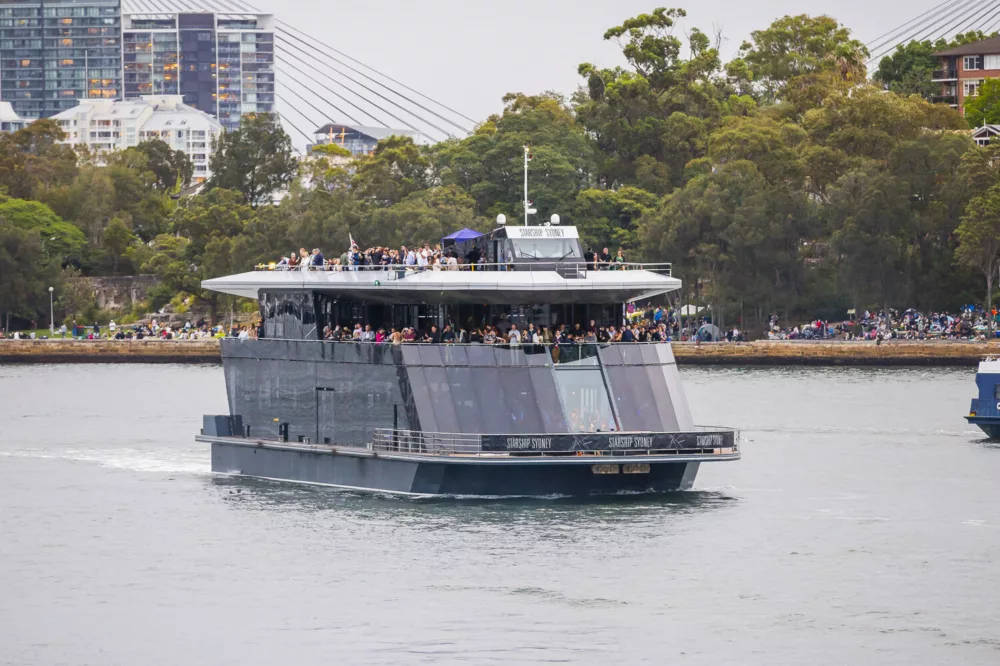 Guests on the top deck of Starship Sydney cruising past a crowded harbour foreshore